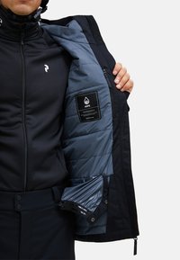 Giacca nera isolante con rivestimento interno blu. Presenta chiusura con zip, imbottitura trapuntata e un'etichetta sulla tasca interna.