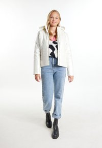 Giacca a vento bianca con cappuccio, caratterizzata da una finitura lucida e due cerniere verticali. Abbinata a un maglione a fantasia bianco e nero e jeans blu.