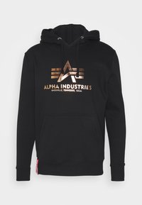 Felpa nera con cappuccio caratterizzata da un logo bronzo con il testo "ALPHA INDUSTRIES". Ha una tasca a marsupio e polsini a costine. Il materiale sembra morbido.