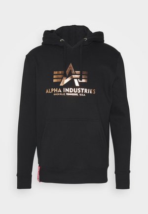 Schwarzer Hoodie mit bronzefarbenem Logo und dem Text "ALPHA INDUSTRIES". Er hat eine Kängurutasche und gerippte Bündchen. Das Material scheint weich zu sein.