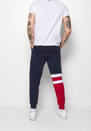 Uomo con le braccia tatuate che indossa una maglietta bianca, pantaloni da jogging blu navy con strisce rosse e bianche su una gamba e scarpe da ginnastica bianche, in piedi di spalle.