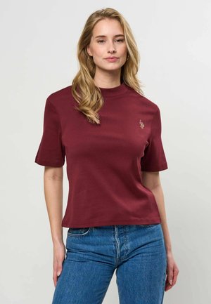 Žena s dlhými blond vlasmi, oblečená v bordovom tričku s krátkym rukávom a modrých džínsoch, stojaca pred svetlosivým pozadím.