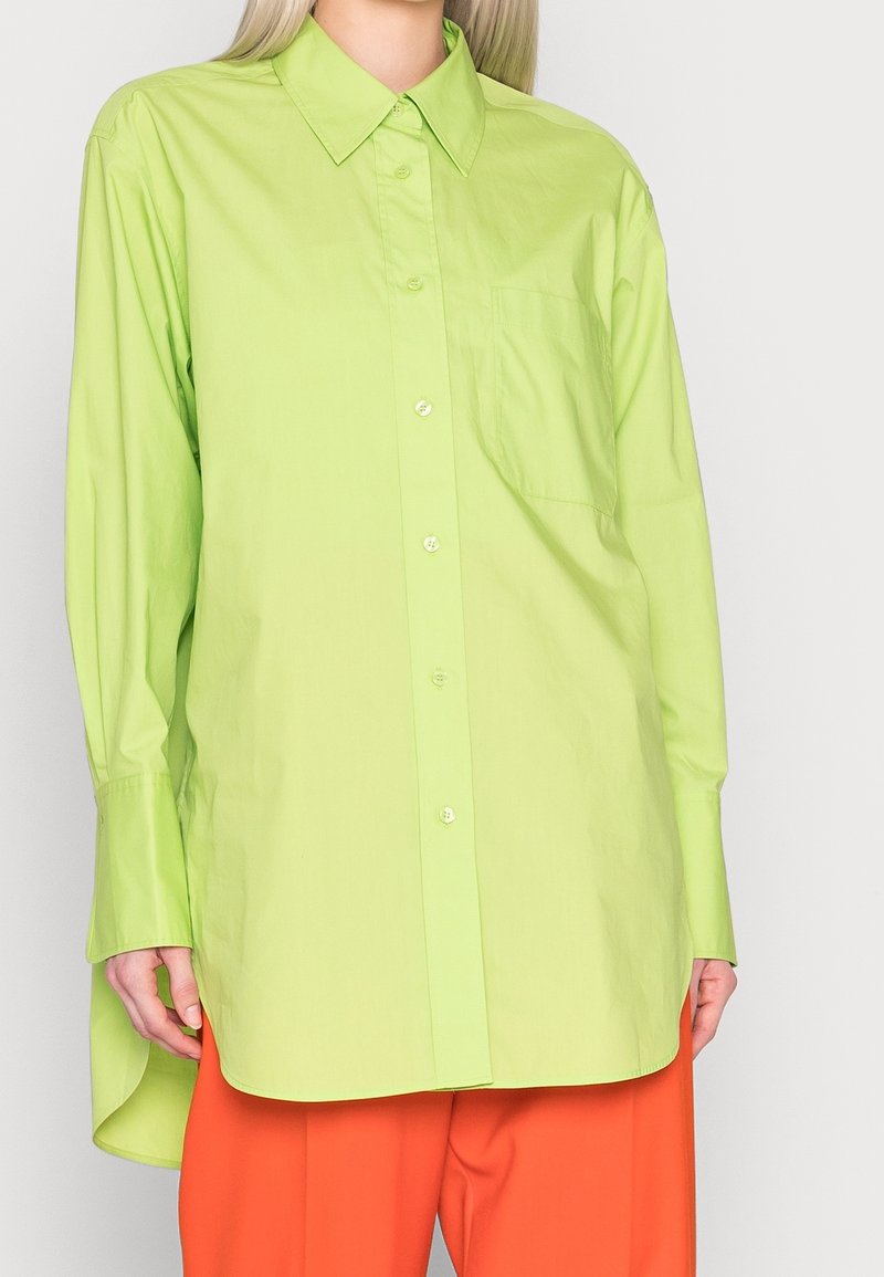 Femme portant une chemise oversize vert citron avec une poche avant et des manches longues, associée à un pantalon orange ample.