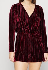 Vínový sametový overal s dlouhými rukávy, hlubokým výstřihem do V, zúženým pasem a šortkami. Má texturovaný vertikální proužkovaný vzor.