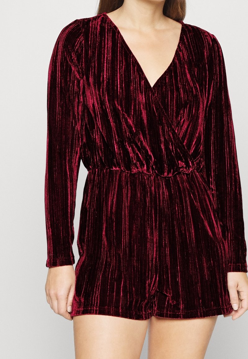 Vínový sametový overal s dlouhými rukávy, hlubokým výstřihem do V, zúženým pasem a šortkami. Má texturovaný vertikální proužkovaný vzor.