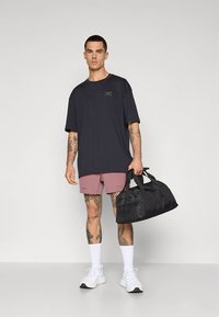 Svart duffelbag med korte håndtak, iført en mørk t-skjorte og rosa shorts. Grå joggesko og hvite sokker, stående på en ensfarget bakgrunn.