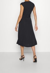 Černé šaty bez rukávů s upnutým korzetem a rozšířenou sukní, s páskem v pase a hladkým, splývavým materiálem. Kombinováno s sandály na vysokém podpatku.