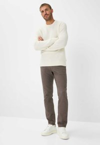 Weißer strukturierter Strickpullover, braune eng geschnittene Hose und weiße Sneakers. Das Model steht mit verschränkten Armen vor einem schlichten weißen Hintergrund.