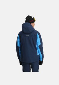 Marineblaue Skijacke mit hellblauen Akzenten, mit Kapuze, Spyder-Logo, langen Ärmeln und strukturiertem Stoff. Handschuhe, die das Modell trägt.