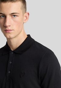 Černé polo tričko s texturovanou látkou, s prošíváním na třech knoflících a vyšívaným logem na levé části hrudníku, disponující klasickým designem límce.