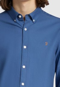 Camicia azzurra con colletto classico, bottoni bianchi e logo "F" arancione sul petto. Tessuto liscio con una leggera lucentezza.