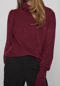 Femme portant un pull à col roulé côtelé bordeaux à manches longues, associé à un pantalon noir, les cheveux lâchés sur l'épaule.