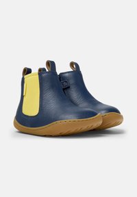 Bottines en cuir bleu marine pour enfants avec des panneaux élastiques latéraux jaunes et des semelles en caoutchouc marron clair, reposant sur un fond blanc.