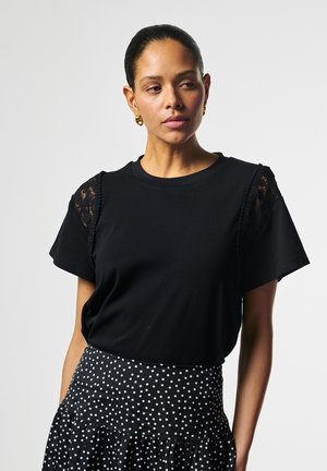 Vrouw met donker haar dat naar achteren is gebonden, draagt een zwart kort mouwtopje met kantaccenten en een zwarte rok met witte stippen, kijkt opzij.