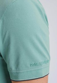 Mintgrön kortärmad polotröja med texturerad tyg. Har en ribbad krage, logotyp på ärmen och kontrasterande sömdetaljer.