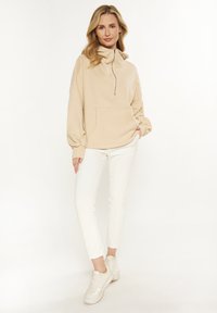 Béžový zip-up pulovr s přední kapsou, uvolněný střih a měkká textura, v kombinaci s bílými slim-fit kalhotami a světlými teniskami.