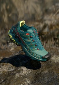 Turkizni trail tekaški čevlj z črnimi in rumenimi akcenti, ki ima teksturirano zgornji del, izstopajoč sistem vezalk in gumijasti pokrov prstov na skalnati površini.