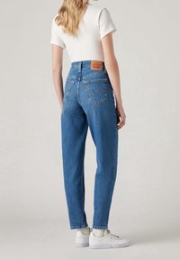 Femme portant un jean en denim bleu taille haute, un haut blanc à manches courtes, des chaussettes blanches et des baskets blanches, debout devant un fond uni.