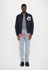 Jachetă bleumarin cu grafic alb și roșu, tricou gri, blugi deschiși cu design roșu și încălțăminte neagră. Îmbrăcăminte casual expusă.
