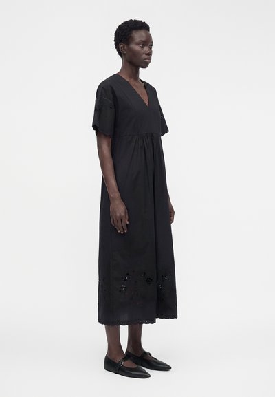 Femme debout dans une robe midi noire à manches courtes avec des motifs floraux découpés près de l'ourlet et sur les bords des manches, portant des ballerines noires.