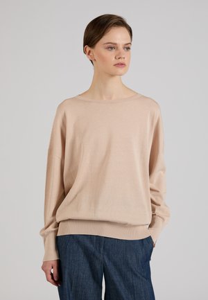 Vrouw met kort bruin haar, die een losse beige trui en blauwe broek draagt, staand tegen een effen lichtgrijze achtergrond.
