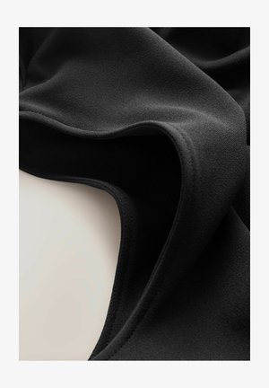 Tessuto nero morbido con bordi lisci e arrotondati e dettagli di cucitura discreti, drappeggiato per mostrare la texture e il movimento su uno sfondo chiaro.