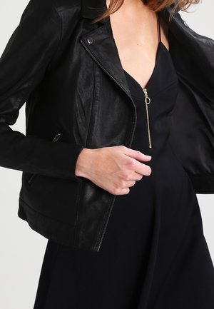 Kobieta ubrana w czarną skórzaną kurtkę na czarnej sukience z złotym zamkiem, trzymająca kurtkę otwartą jedną ręką.