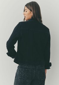 Mustmustest denim-jakk, millel on klassikaline lõike, rullitud varrukad ja tekstuuriga must pükste muster. Lihtne disain koos nähtavate õmbluse detailidega.