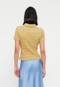 Γυ ναίκα με κοντά μαύρα μαλλιά που φοράει κίτρινη μπλούζα με υφή και κοντά μανίκια και φούστα από γαλάζιο σατέν, εμφανίζεται από πίσω με λευκό φόντο.