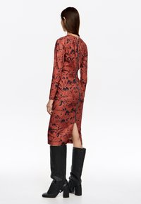 Vestido de manga larga ajustado en óxido con manchas negras, con cremallera en la parte trasera y una abertura en el dobladillo, combinado con botas altas negras.