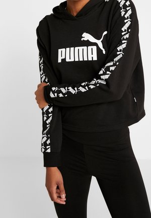 Osoba nosząca czarną bluzę z kapturem Puma z białym logo z przodu i wzorzystymi paskami z logo na obu rękawach, w połączeniu z czarnymi spodniami.