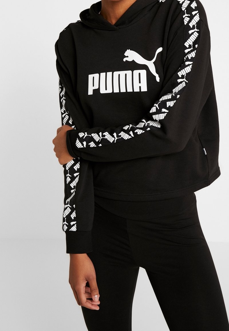 Personne portant un sweat à capuche noir Puma avec un logo blanc sur le devant et des bandes de logo à motifs le long des deux manches, associé à un pantalon noir.