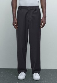 Man in een losse zwarte broek en witte sneakers, staand tegen een lichtgrijze muur op een donker gespikkelde vloer.