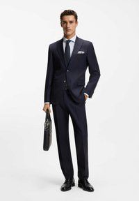 Młody mężczyzna w ciemnym, wąsko prążkowanym garniturze, białej koszuli i krawacie w kropki, trzymający czarną teczkę, stojący na białym tle.