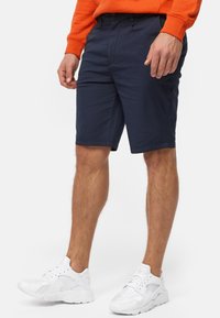 Dunkelblaue Shorts aus Baumwollmischung mit Knopfverschluss, Seitentaschen und geradem Schnitt. Kombiniert mit weißen Sportschuhen.
