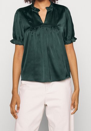 Mørkegrøn satinbluse med flæset korte ærmer og krave, kombineret med lyse bukser på en stående person.