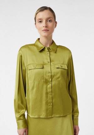 Woman wearing a shiny green button-up shirt with chest pockets, standing against a plain white background.