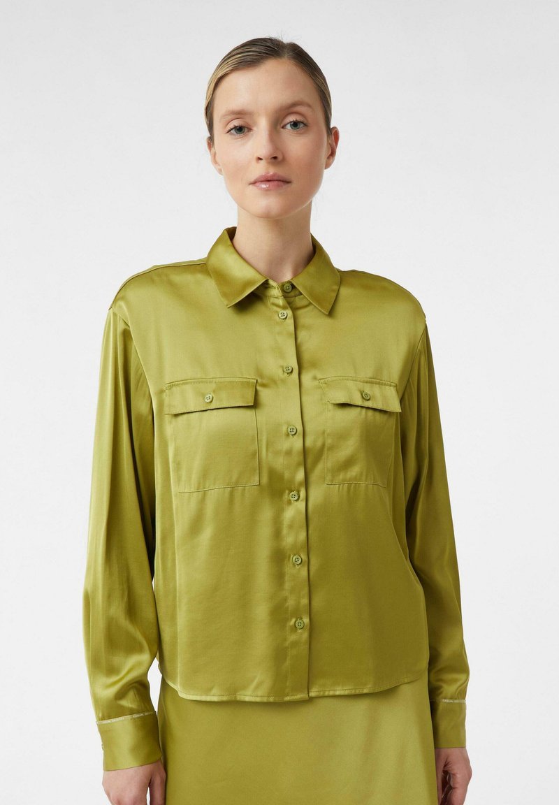Woman wearing a shiny green button-up shirt with chest pockets, standing against a plain white background.