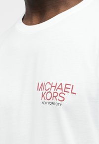 Κοντινό πλάνο ενός ατόμου που φορά λευκό μπλουζάκι με κόκκινο κείμενο "MICHAEL KORS" και μικρότερο μαύρο κείμενο "NEW YORK CITY" στο στήθος.