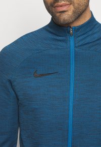 Blaue Sportjacke aus strukturiertem Stoff, mit hohem Kragen, durchgehendem Reißverschluss und schwarzem Nike-Logo auf der Brust.
