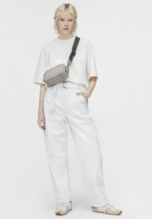 Gray rectangular crossbody bag with "TOUS" embossed, adjustable strap; model wearing a loose white shirt and wide-leg white pants.