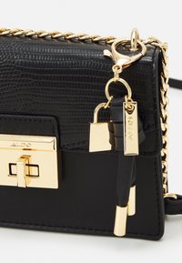 Černá kabelka ze syntetického materiálu s texturou, se zlatými detaily, přední zámkovou funkcí a řetězovými akcenty. Obsahuje dekorativní zámek a štítek.