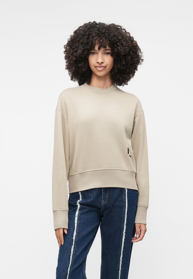 Sweatshirt bej scurt cu mâneci lungi, guler rotund și un buzunar frontal. Asortat cu blugi albaștri cu dungi laterale albe.