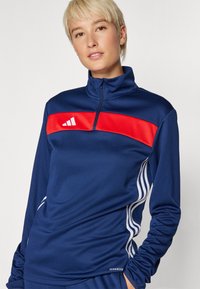 Námornícky atletický pulover s červeným horizontálnym pruhom, zipsom na krku, bielymi bočnými pruhmi a detailom textu "AEROREADY" na okraji. Mäkkučký materiál.