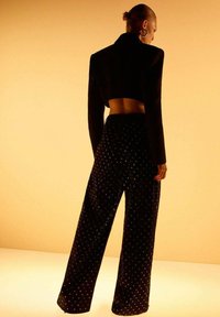 Mujer con el cabello recogido en un moño, viste una chaqueta corta negra y pantalones de pierna ancha con un patrón de puntos brillantes, de pie contra un fondo amarillo cálido.