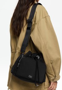 Čierna nylonová taška s pevne stiahnutým vrchom, zipsovým vreckom a nastaviteľným popruhom. Obsahuje kovové akcenty a detaily s logom.