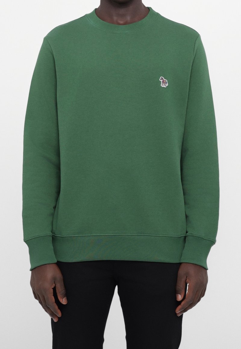 Homme portant un sweat-shirt vert uni à col rond avec un petit logo zèbre multicolore brodé sur le côté gauche de la poitrine, associé à un pantalon noir.