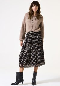 Frau steht und trägt einen taubfarbenen Schnürpullover, einen schwarz-beige gemusterten, plissierten Midirock, einen schwarzen Gürtel und schwarze spitz zulaufende Mid-Heel-Stiefel.