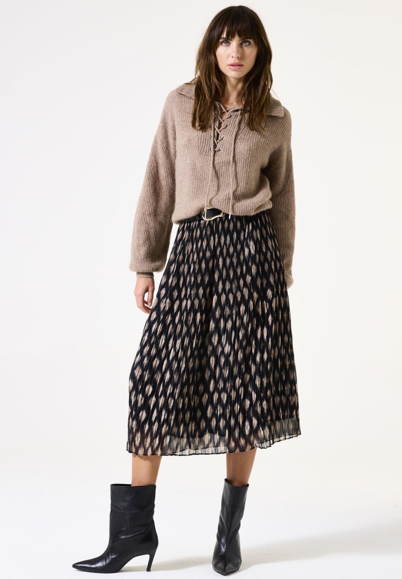 Frau steht und trägt einen taubfarbenen Schnürpullover, einen schwarz-beige gemusterten, plissierten Midirock, einen schwarzen Gürtel und schwarze spitz zulaufende Mid-Heel-Stiefel.