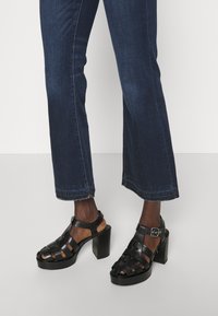 Sandalias de tacón negro con un diseño trenzado, punta abierta y tira en el tobillo. Combinadas con unos jeans recortados de color azul oscuro con bordes deshilachados.